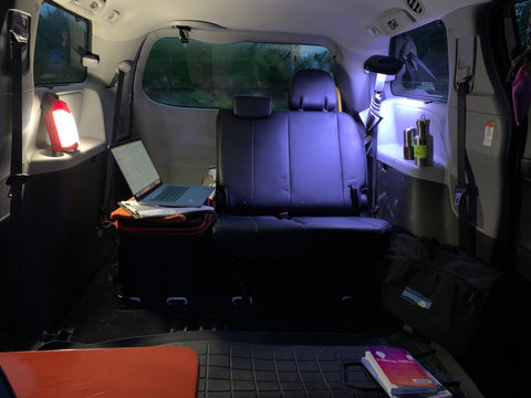 The inside of a mini van with the seats pulled out, lit by camping lanterns with a laptop, and a mobile office desk