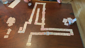 Video of a woman shuffling the quarter-size playing cards "Monaco-style". The mini cards are splayed out on the table face-up from a game. She scoops them towards herself, flips them over in one mass, and then moves them in circles around the table to shuffle. 