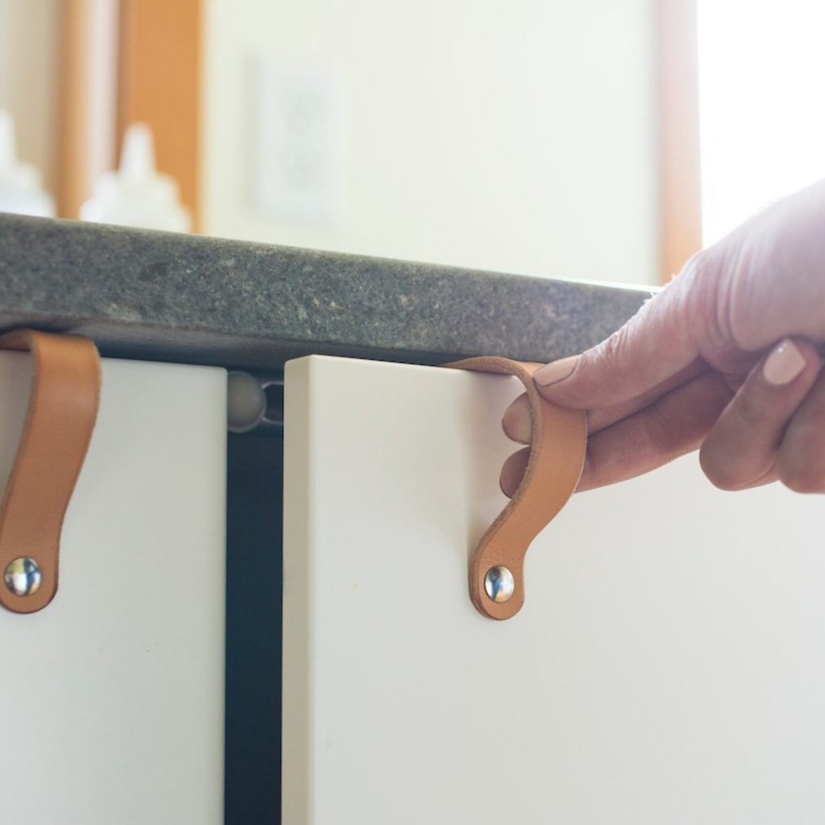 Woman is pulling open a bottom cabinet door under sink with a natural leather Lovejoy handle. On top of the cabinet is the two tiny wood screws that attach the handle to the IKEA cabinet door.