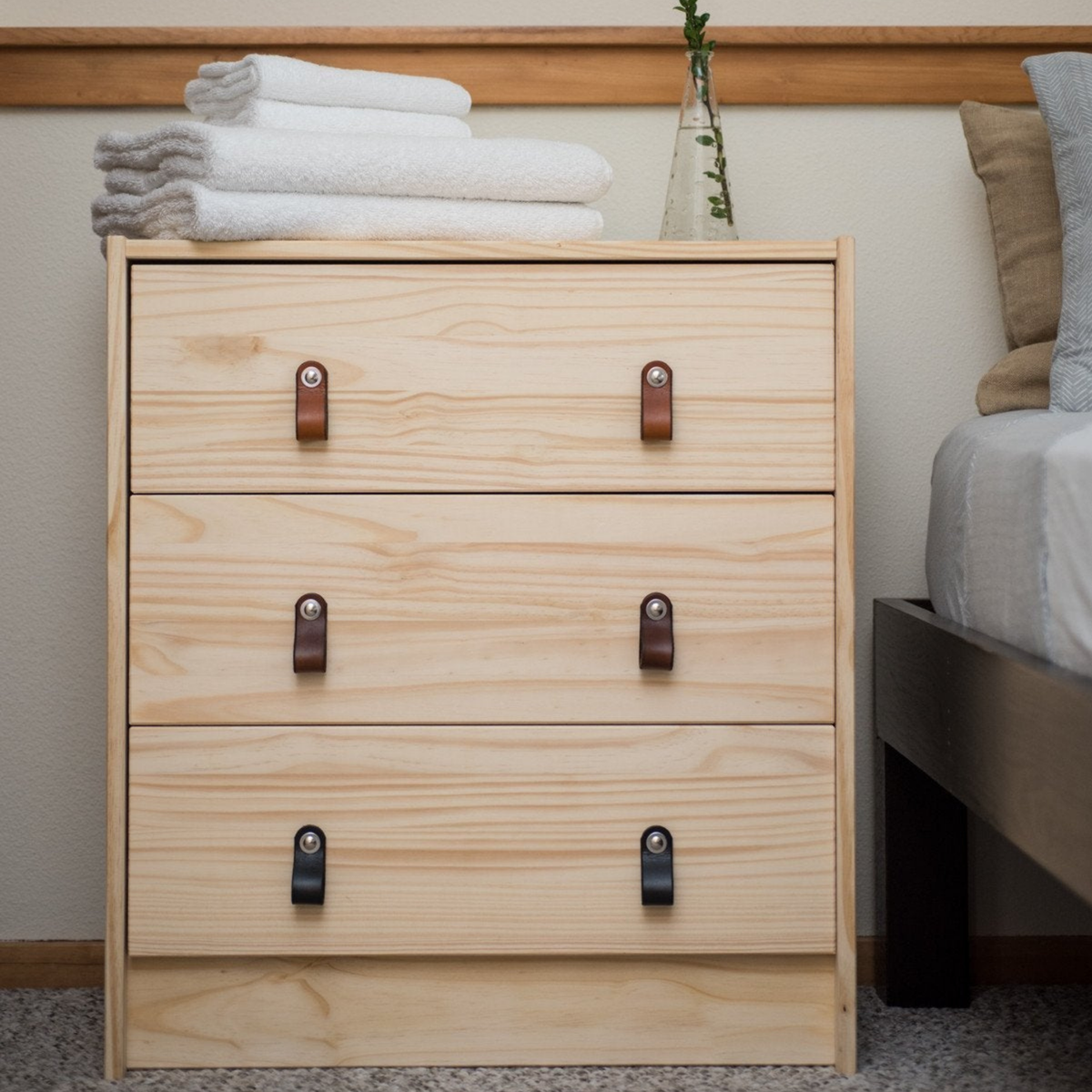 Inexpensive knotty pine dresser from IKEA seen from across the room. Each drawer has two small leather Hawthorne pulls in a different color, arranged from light to dark: honey, dark brown, and black. 