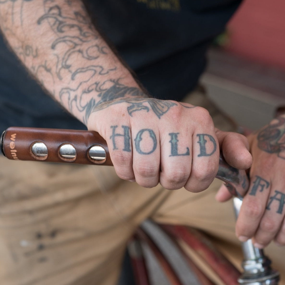 Tattooed man resting his hand on a bicycle city bar wrapped in a dark brown leather grip that has &quot;porthole&quot; round cutouts for finger holds. The man&#39;s hand tattoos say &quot;HOLD FAST&quot;