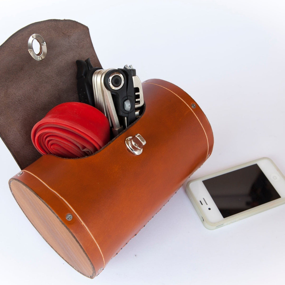 Honey leather variant of leather bicycle barrel bag on a plain white background. Inside the bag is a multi-tool and spare bike tube. An iPhone 4 is beside the bag for scale.