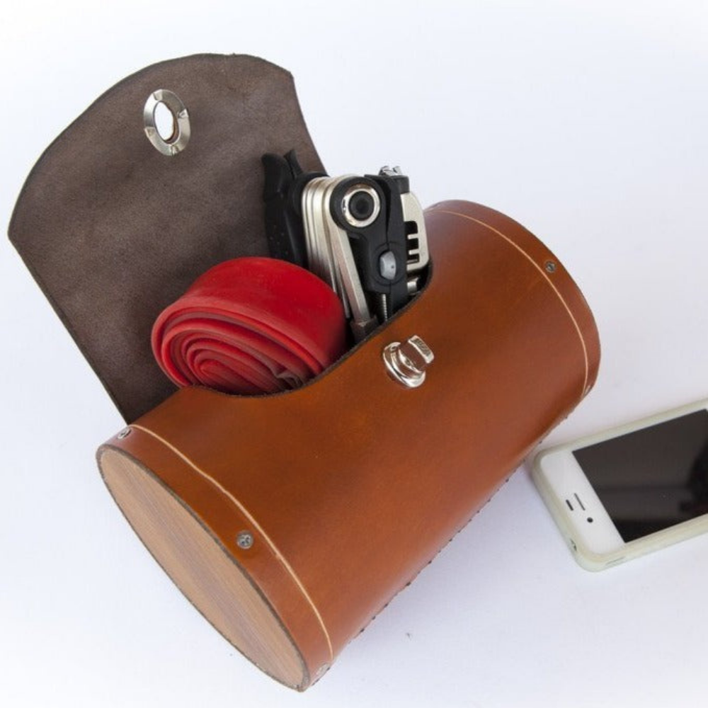 Honey leather variant of leather bicycle barrel bag on a plain white background. Inside the bag is a multi-tool and spare bike tube. An iPhone 4 is beside the bag for scale.