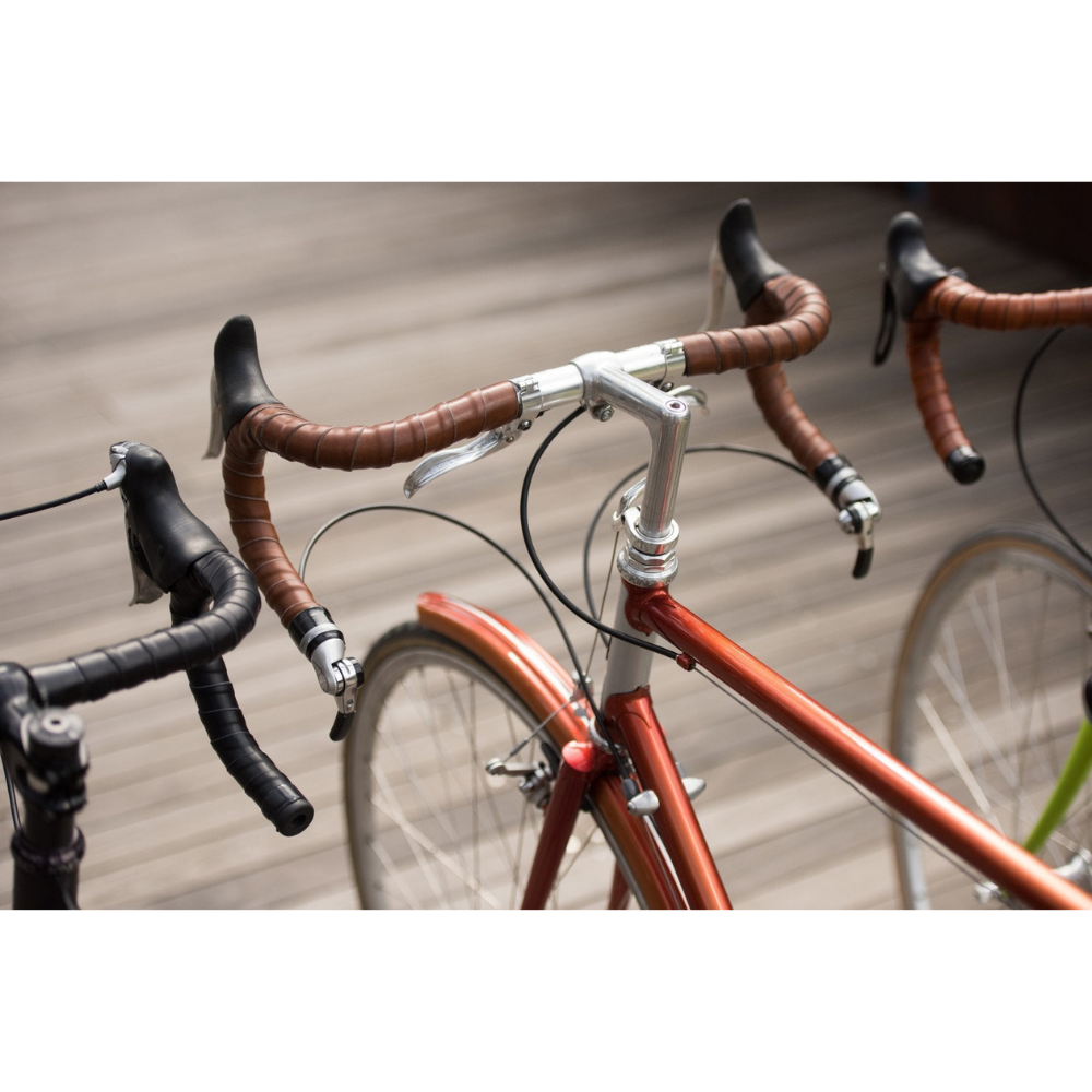 A bird&#39;s eye view looking down on three bicycles lined up in a row over a boardwalk floor. Each bicycle has a different color leather wrapped in coils on its handlebars: Dark Brown, Black and Honey color.