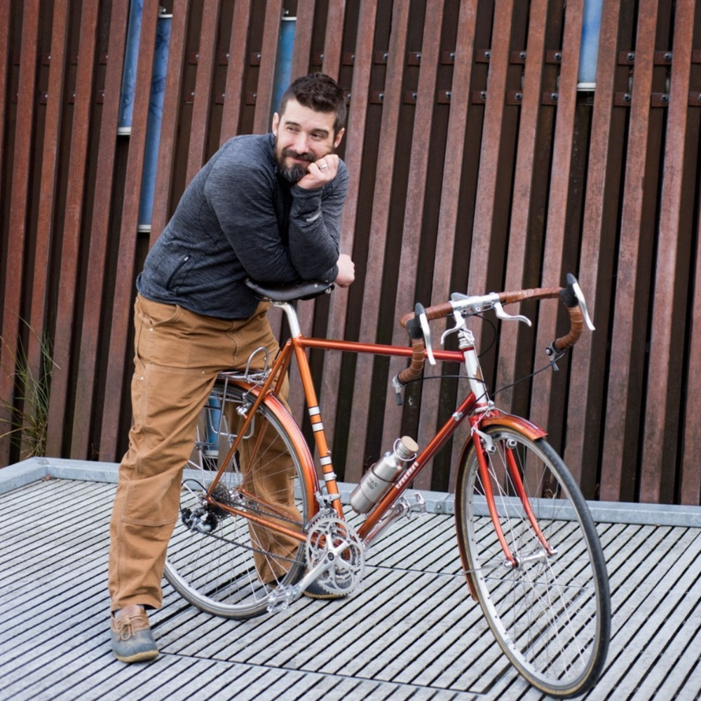 Orange steel frame road bicycle with a man standing behind it, his legs straddling the back wheel. He is resting his elbow on the saddle and has a wistful half-smile. The handlebars are wrapped in coiled dark brown vegetable-tanned leather. 