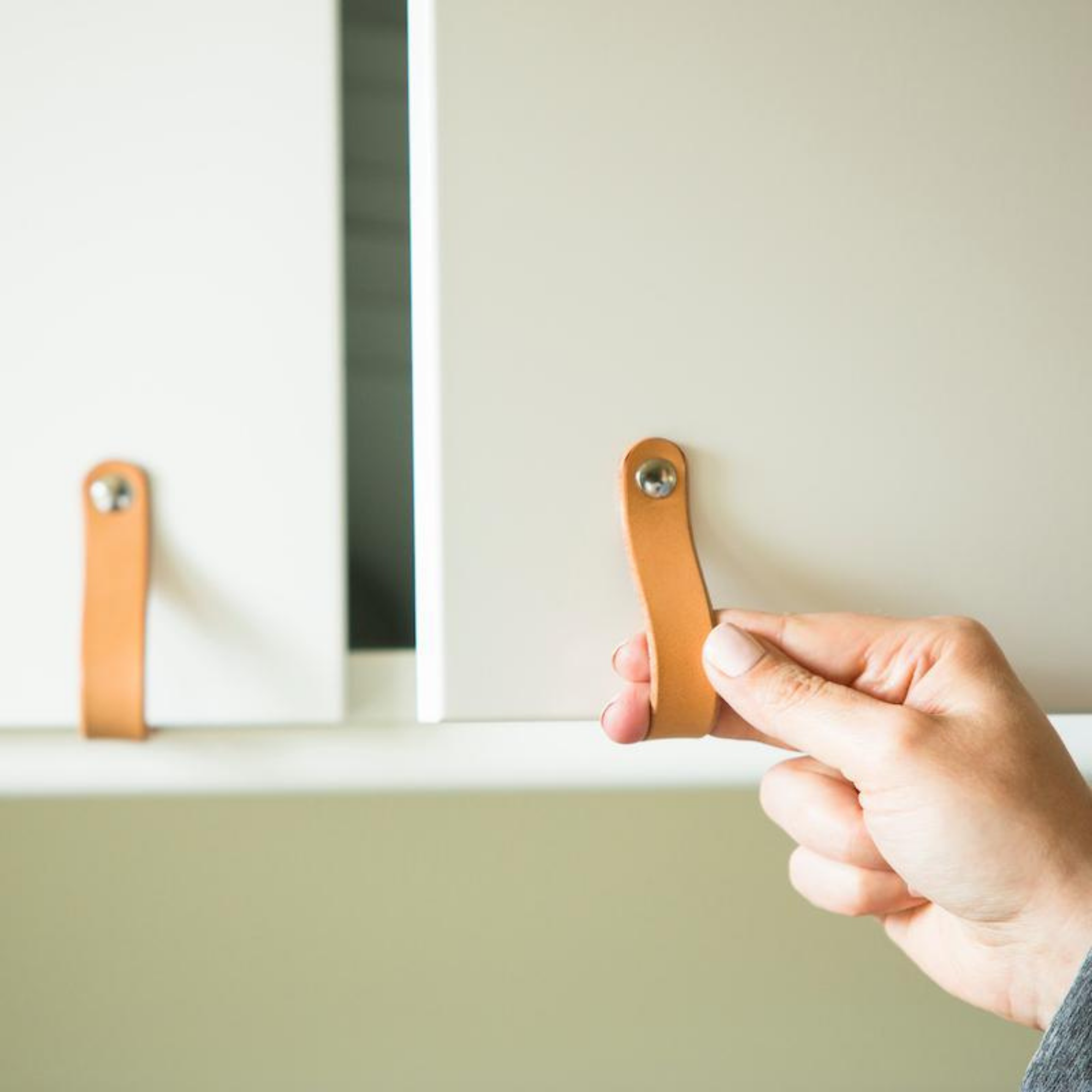 A woman's hand is reaching towards a tan, natural leather cabinet handle on a set of white cabinets. The handle loops around the bottom of the European style (IKEA) cabinet door and attaches on the underside.