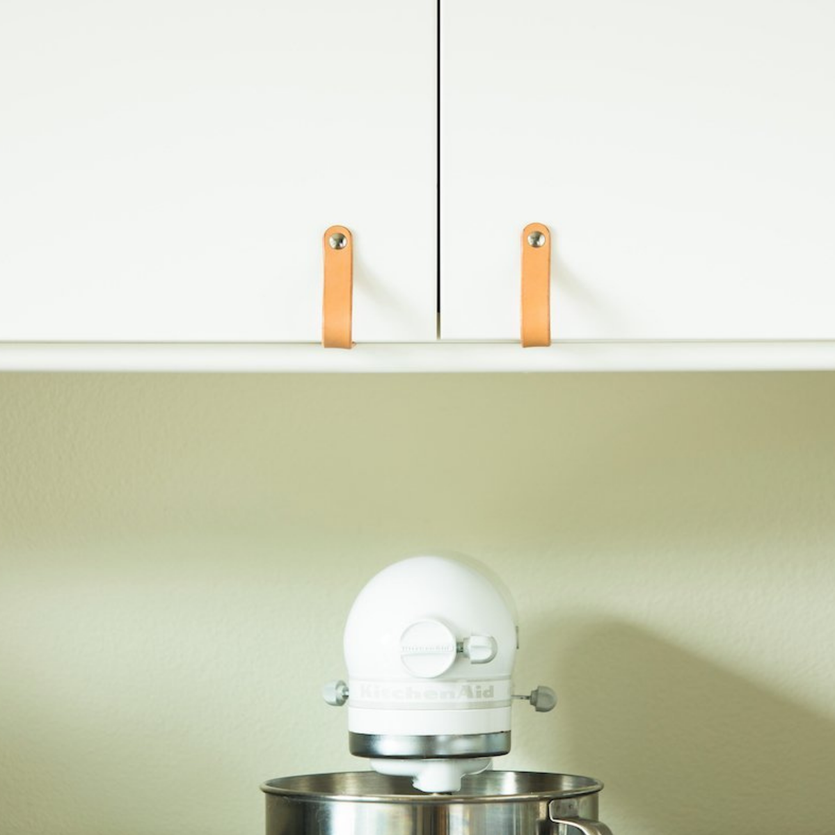 White kitchen with white walls, white cabinets, and a white Kitchenaid mixer on the counter with soothing tan natural vegetable-tanned leather Lovejoy handles on the cabinet doors. 