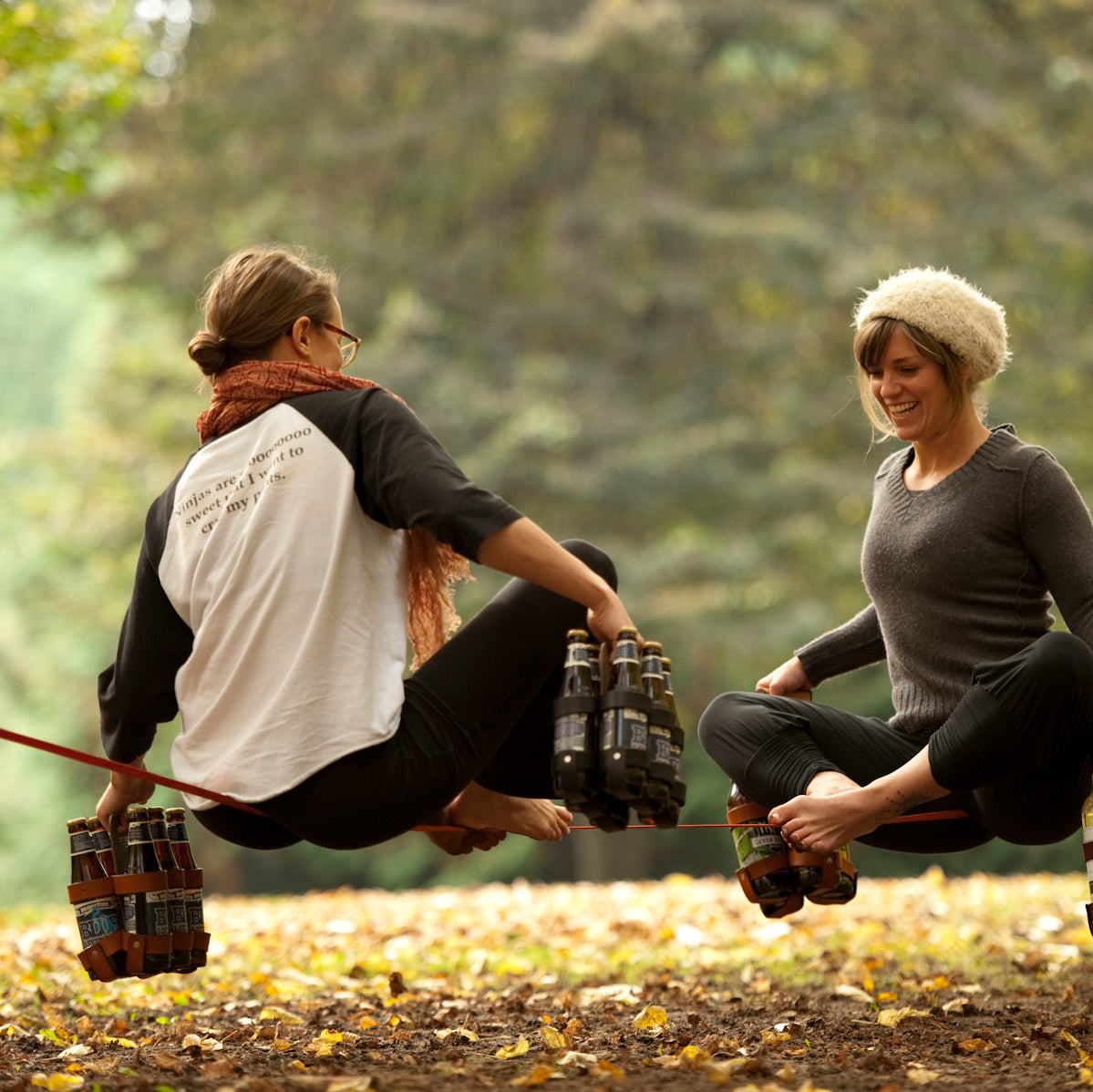 Two women balancing on a slackline in a park in Portland Oregon brewery town, holding leather 6-packs of beer as balance weights